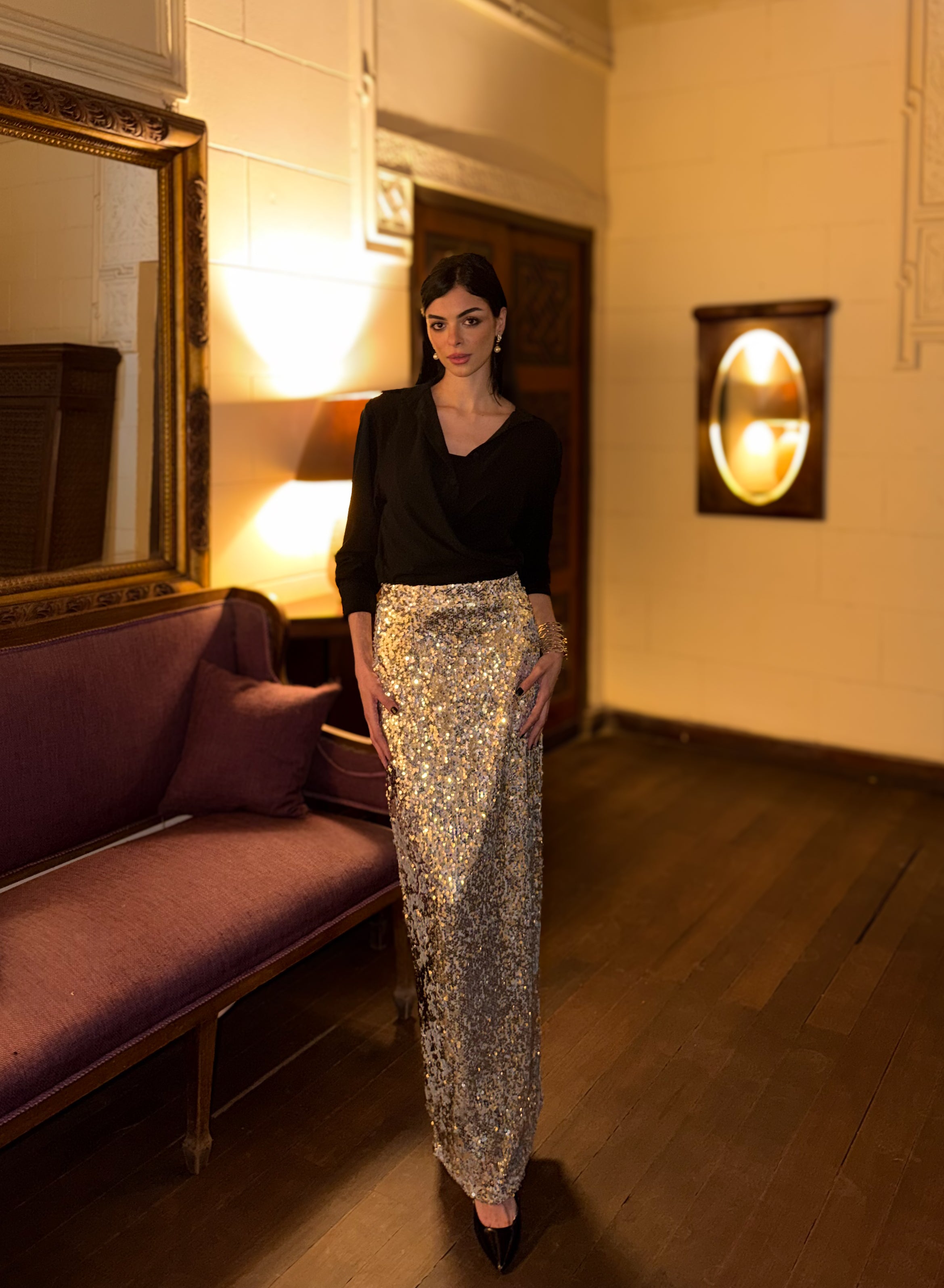 Woman in a black top and sequin skirt standing in a room with a purple couch and mirror.